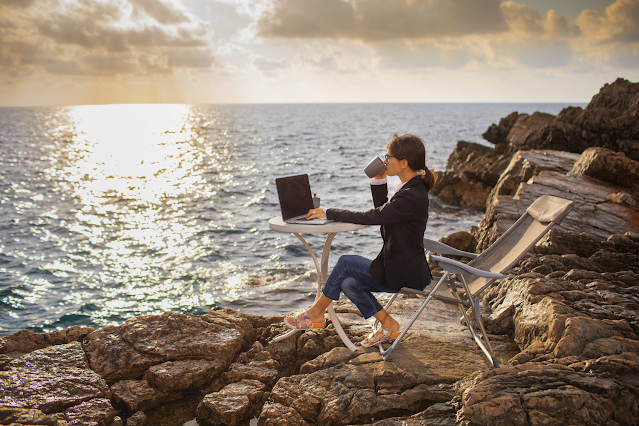 A lady seated on a chair by a pebbly shoreline, enjoying a cup of coffee and working on a laptop.